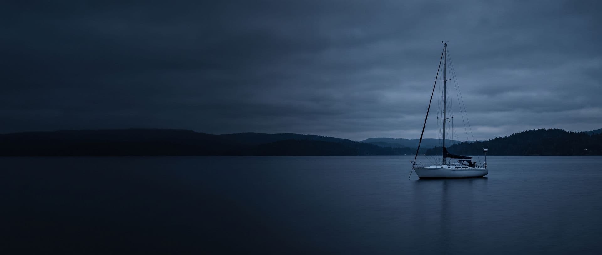 A sailboat lying at anchor on flat open water under a low overcast sky at dusk