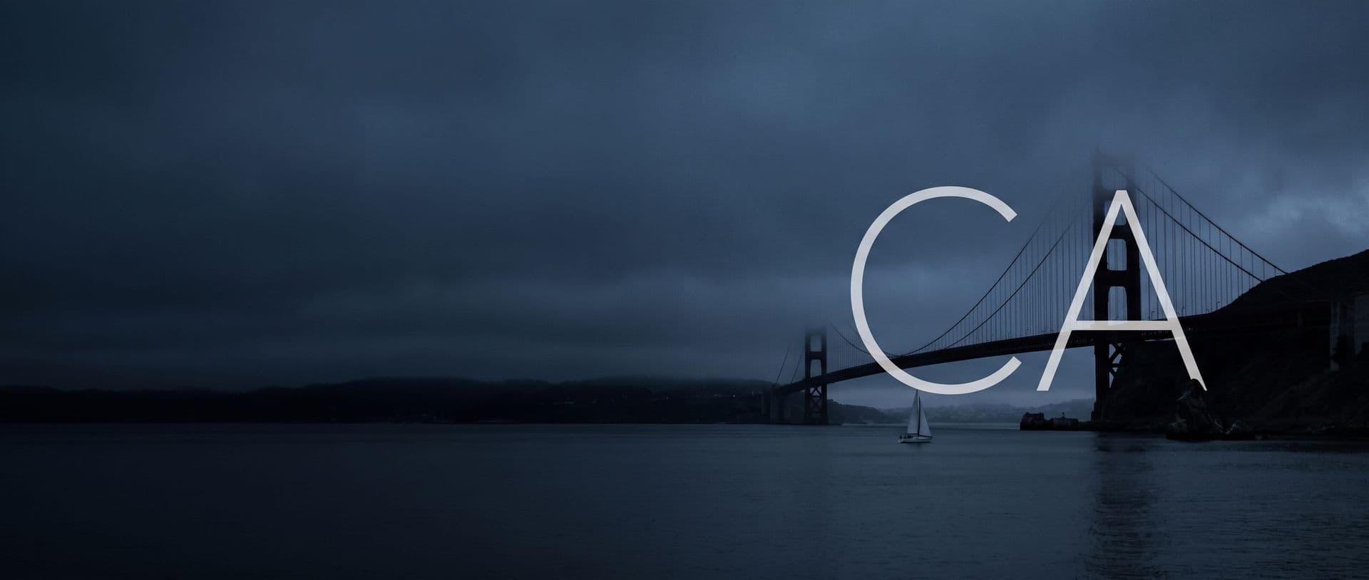 The Golden Gate Bridge in fog at dusk, a sailboat passing under the span, with the letters CA in the sky