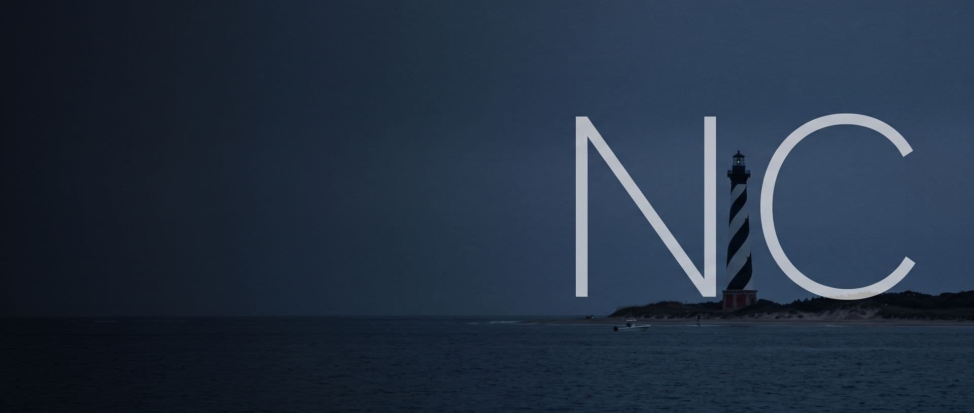 The Cape Hatteras lighthouse on the Outer Banks at dusk, a boat offshore, with the letters NC in the sky