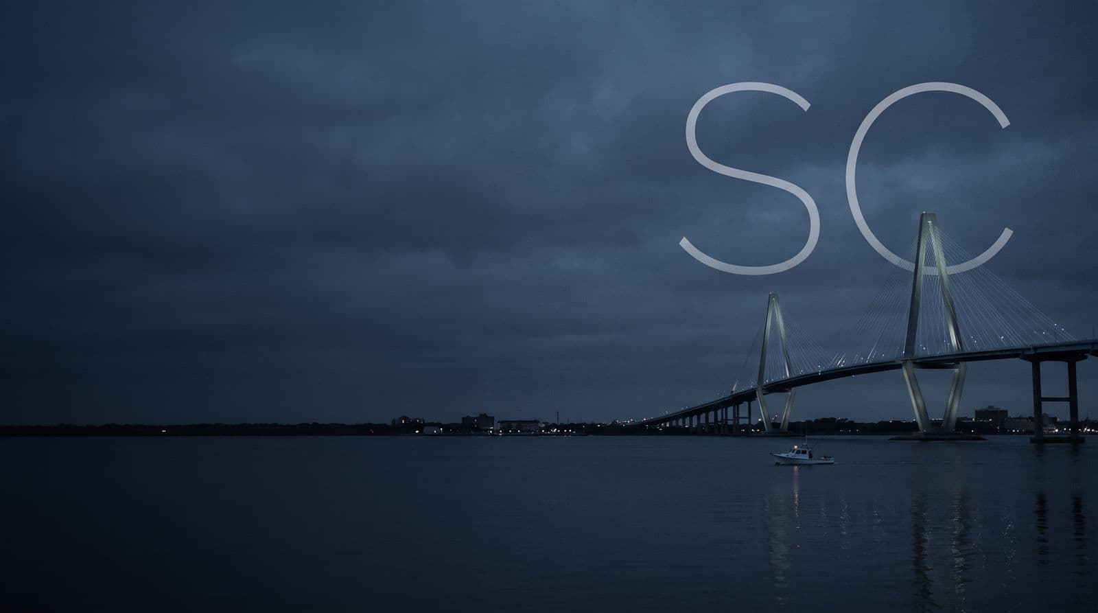 The Ravenel Bridge over Charleston harbor at dusk, a boat on the water below, with the letters SC in the sky