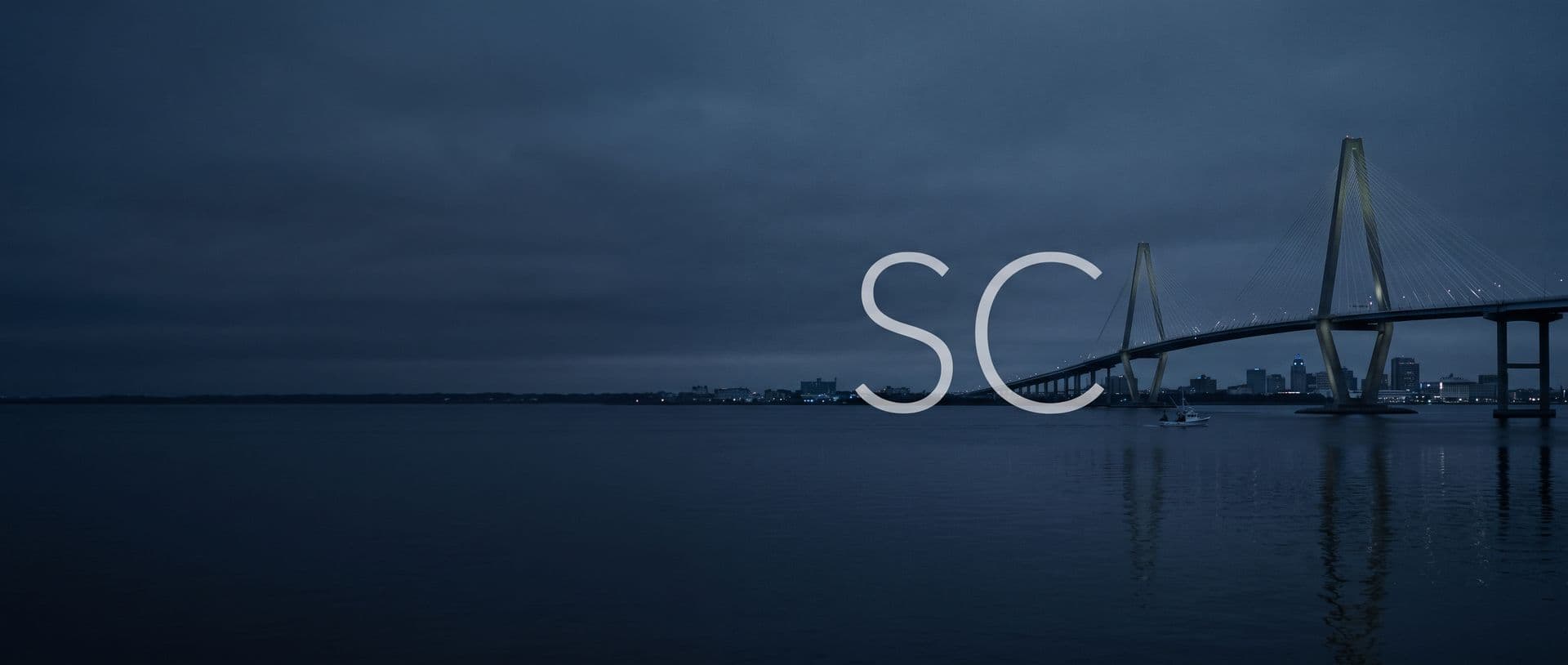 The Ravenel Bridge over Charleston harbor at dusk, a boat on the water below, with the letters SC in the sky