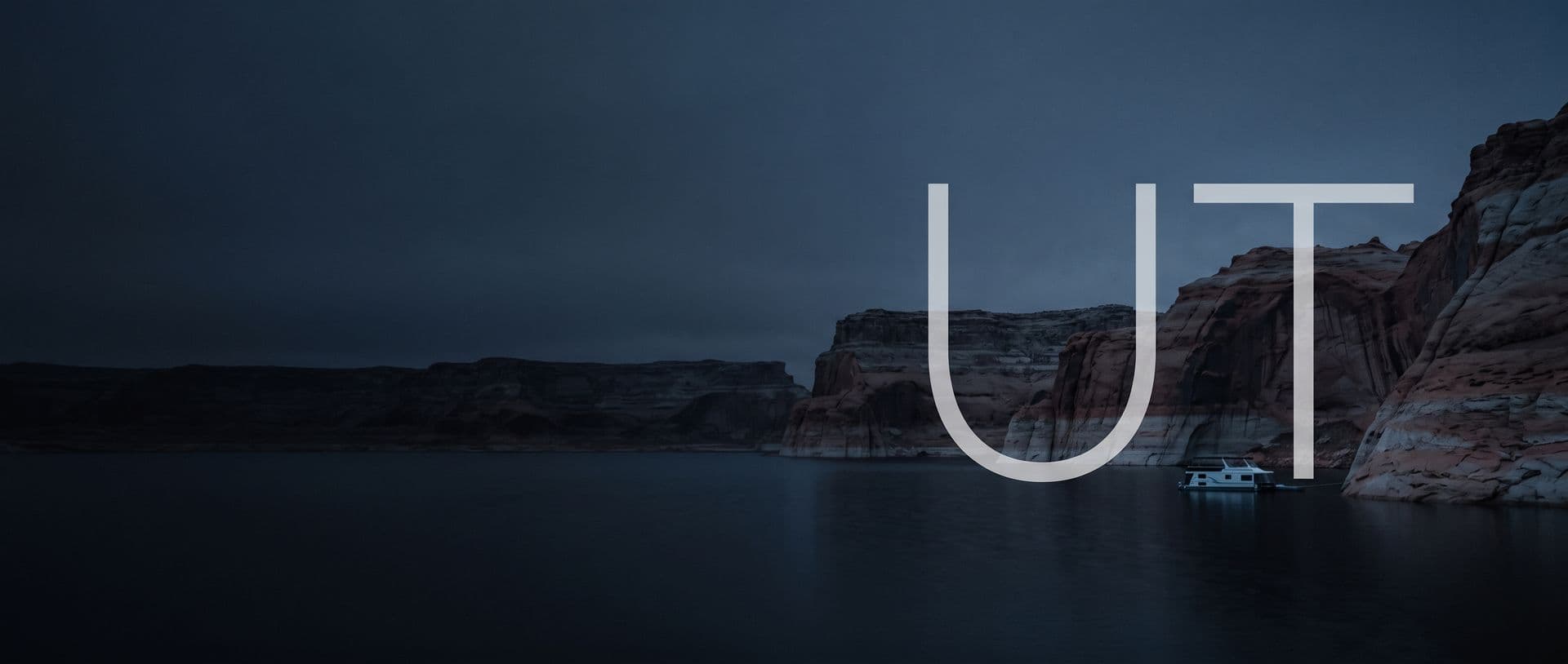 Sandstone canyon walls on Lake Powell at dusk, a houseboat moored in their shadow, with the letters UT in the sky