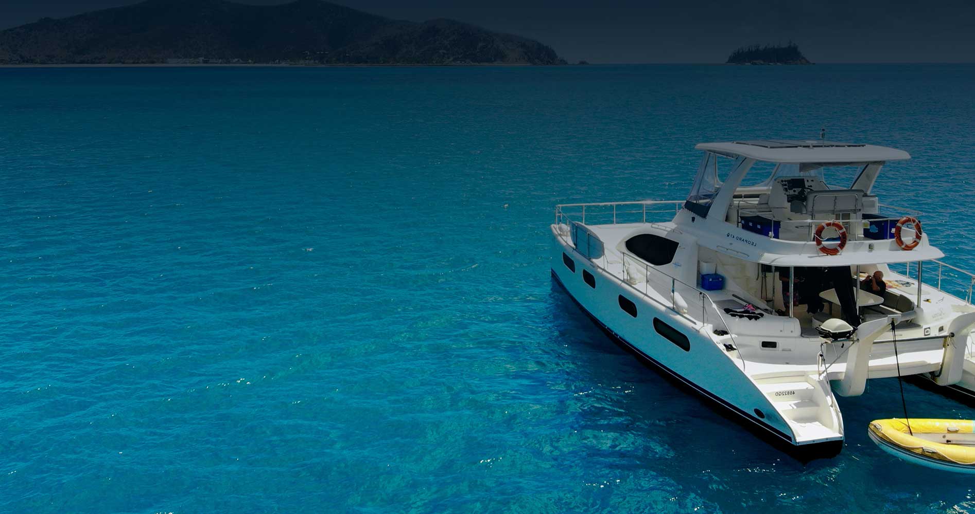 Power catamaran lying at anchor over bright turquoise shallows, with dark headlands on the horizon behind her