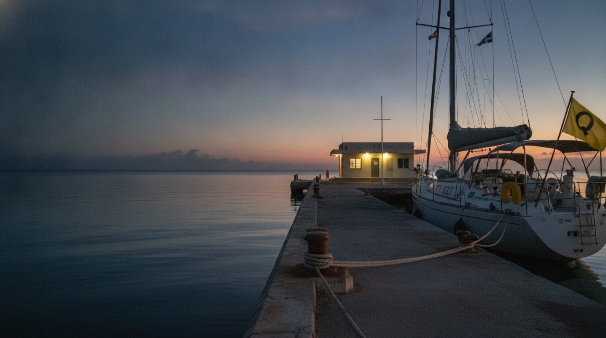 A cruising yacht lying to a courtesy flag off a customs dock at first light, waiting to clear in
