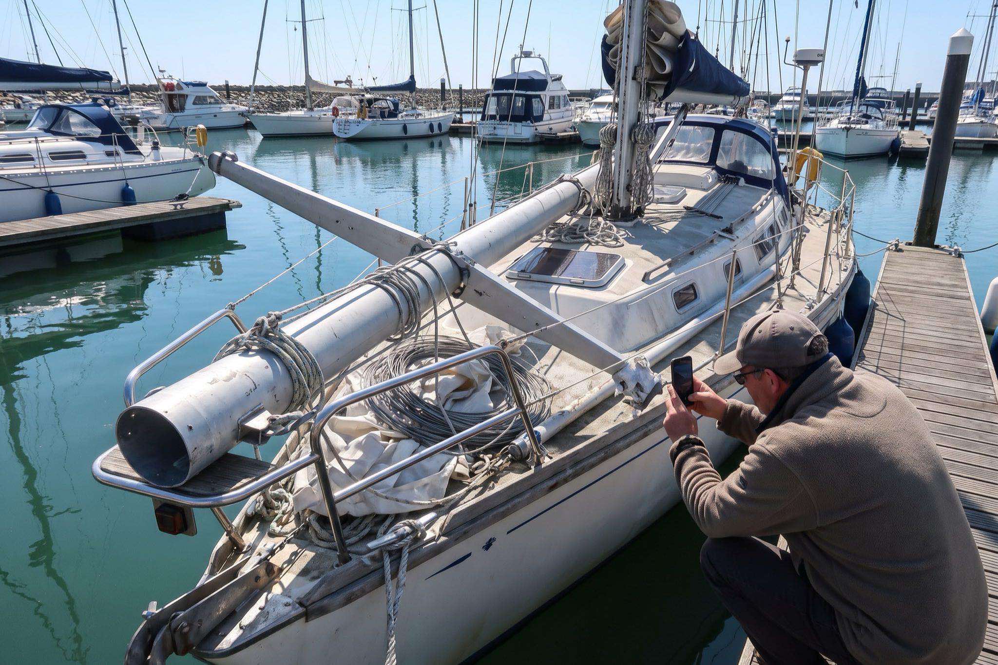 A dismasted cruising yacht alongside a marina pontoon, the broken mast lashed on deck while the owner photographs the failed fitting
