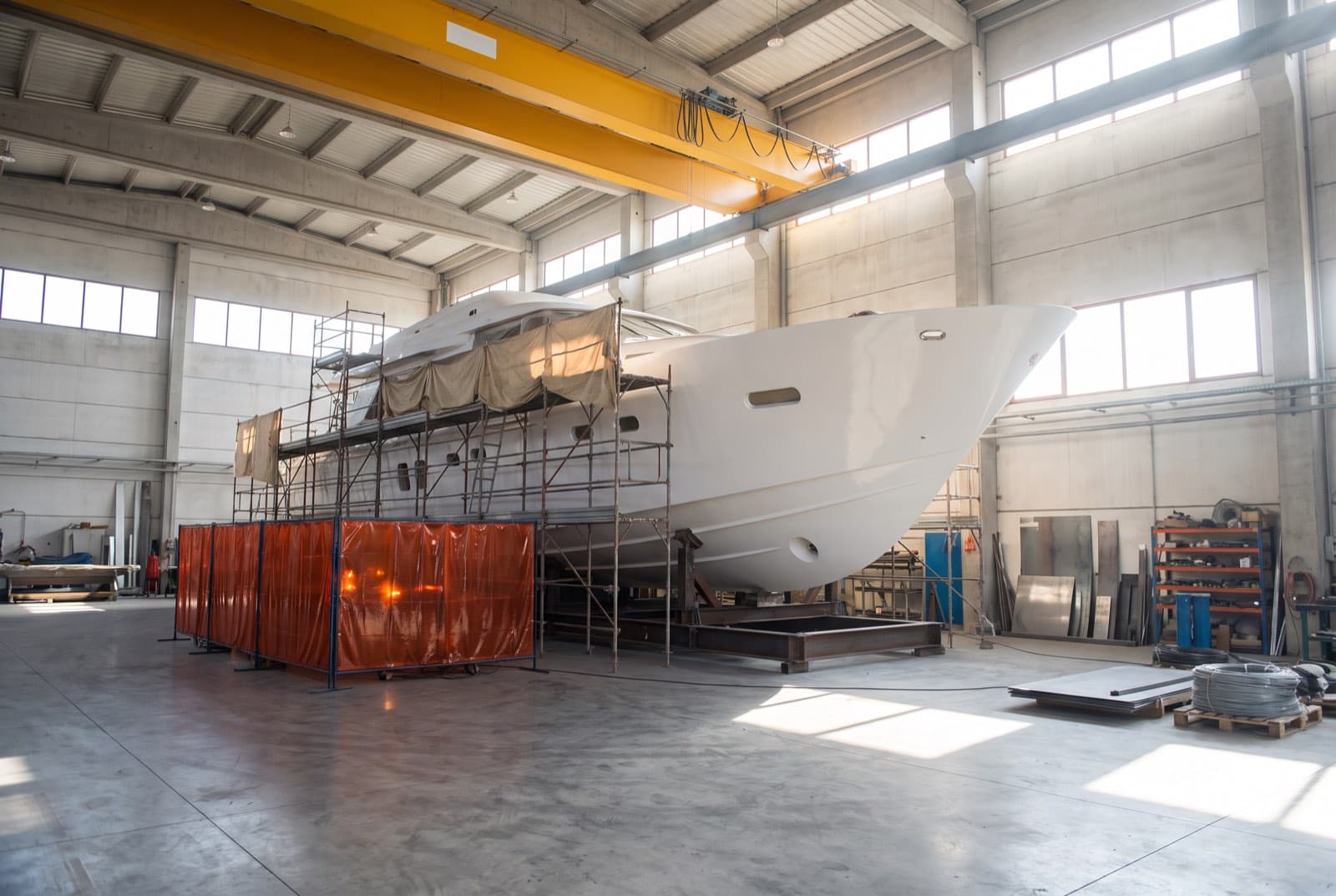 A white motor yacht hull on cradles inside a bright yacht construction hall, with scaffolding along the topsides and orange welding screens by the hull