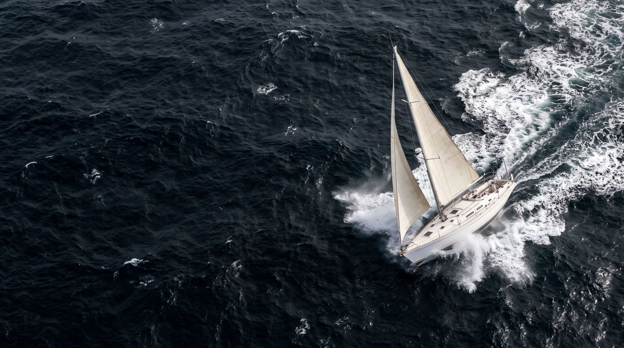 Aerial view of a white sailing yacht heeled hard over in heavy breaking seas during a daytime storm, the weather a hurricane plan is written to keep a boat out of
