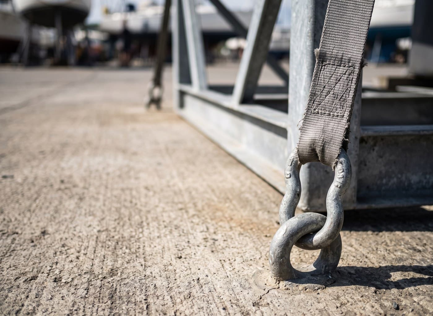 Close daylight detail of a yacht cradle and galvanised tie-down strap shackled to an embedded concrete ground anchor on a boatyard hardstanding
