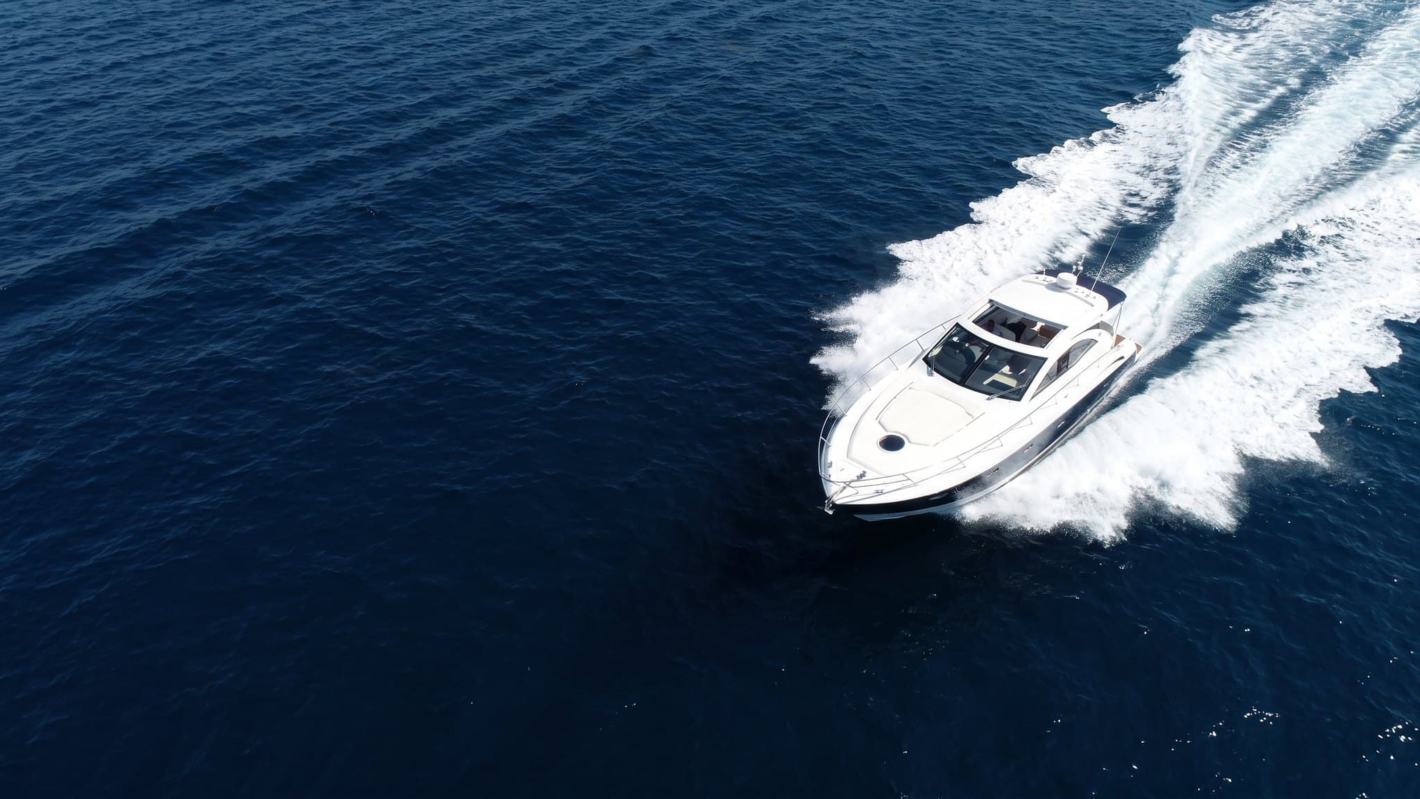 White and navy sports powerboat running at speed across deep blue open water in bright midday sun, seen from a high oblique aerial with a broad white wake behind it