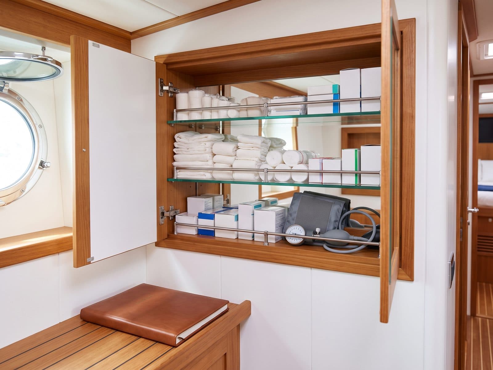 An open medical cabinet with bandages, dressings and a blood pressure cuff in the crew area of a yacht, a porthole on the left