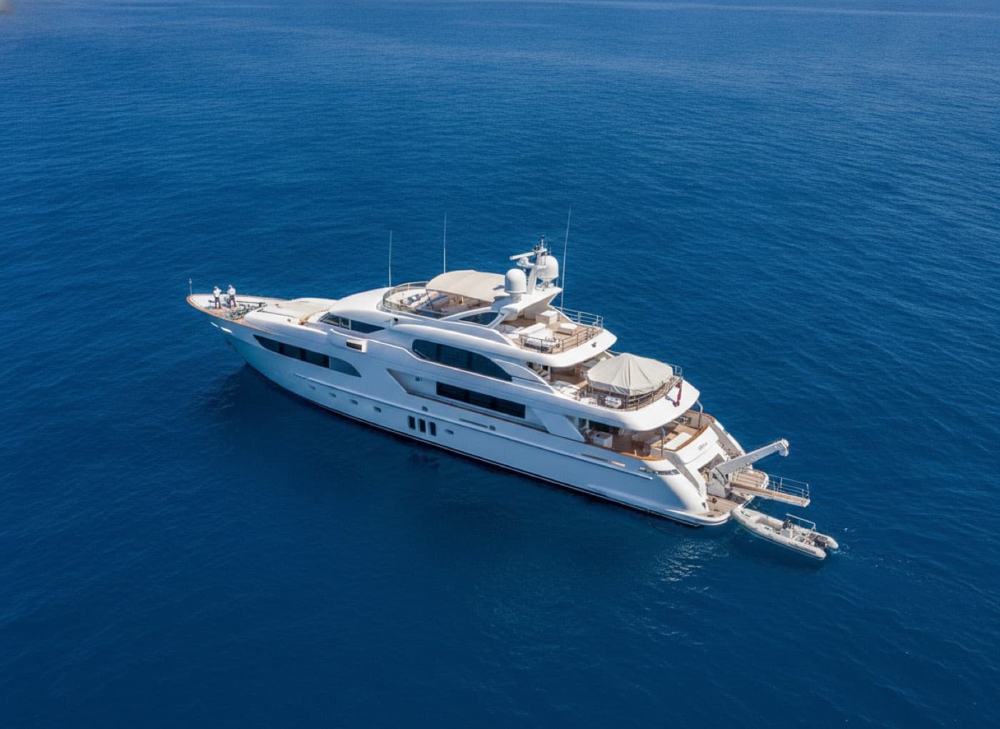 A large crewed motor yacht lying at anchor in deep blue open water under bright midday sun, two crew working on the foredeck and a tender alongside