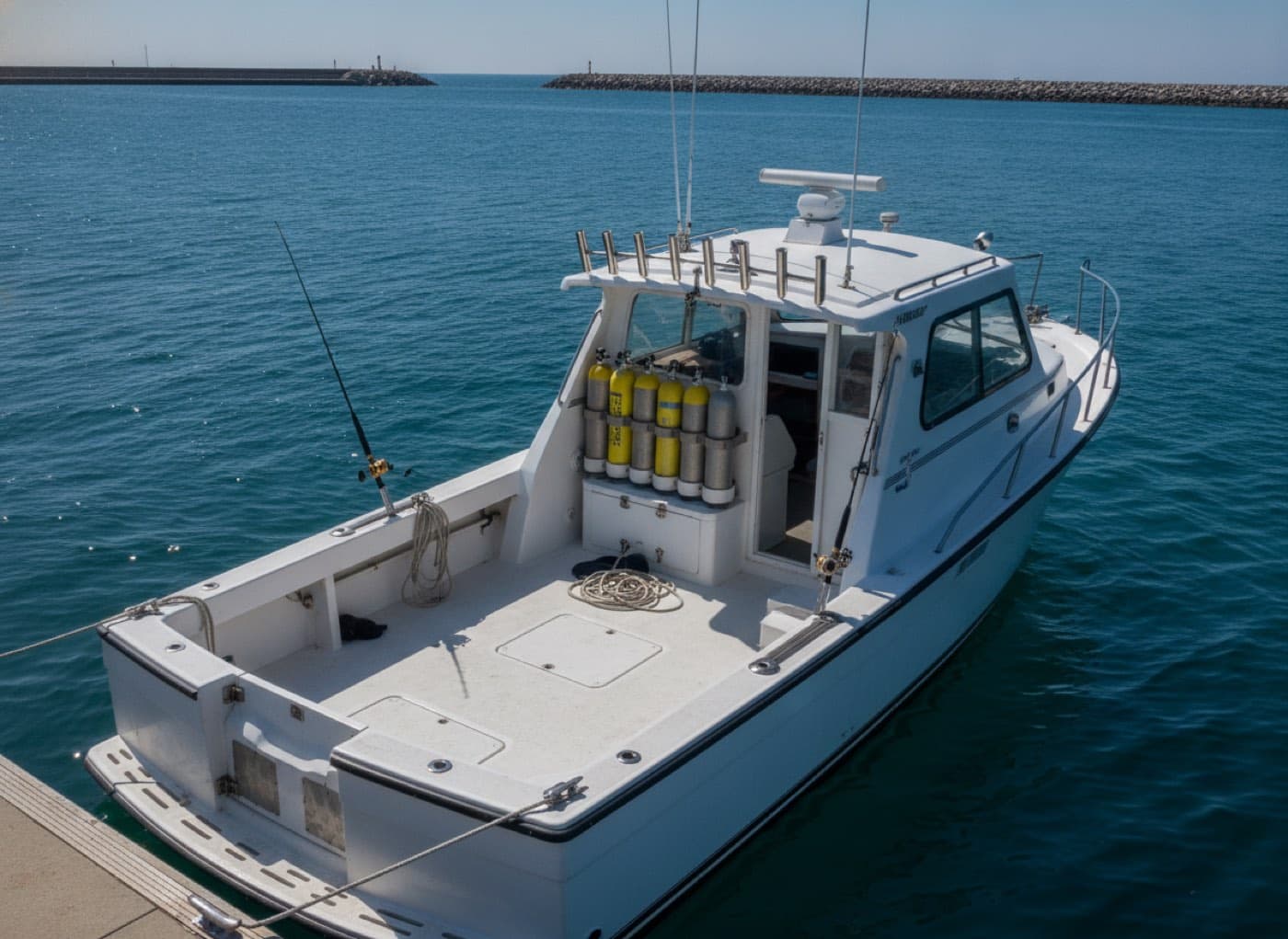A 38-foot dive and angling charter boat moored at a sunlit pontoon, scuba tanks racked on deck and rod holders along the hardtop, deep blue water behind