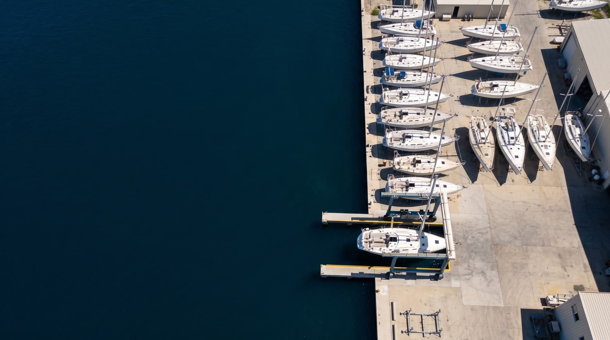 High oblique aerial view of a Caribbean boatyard in bright midday sun, sailing yachts standing on cradles beside a travel-lift dock with deep blue open water alongside, the two places a boat spends hurricane season