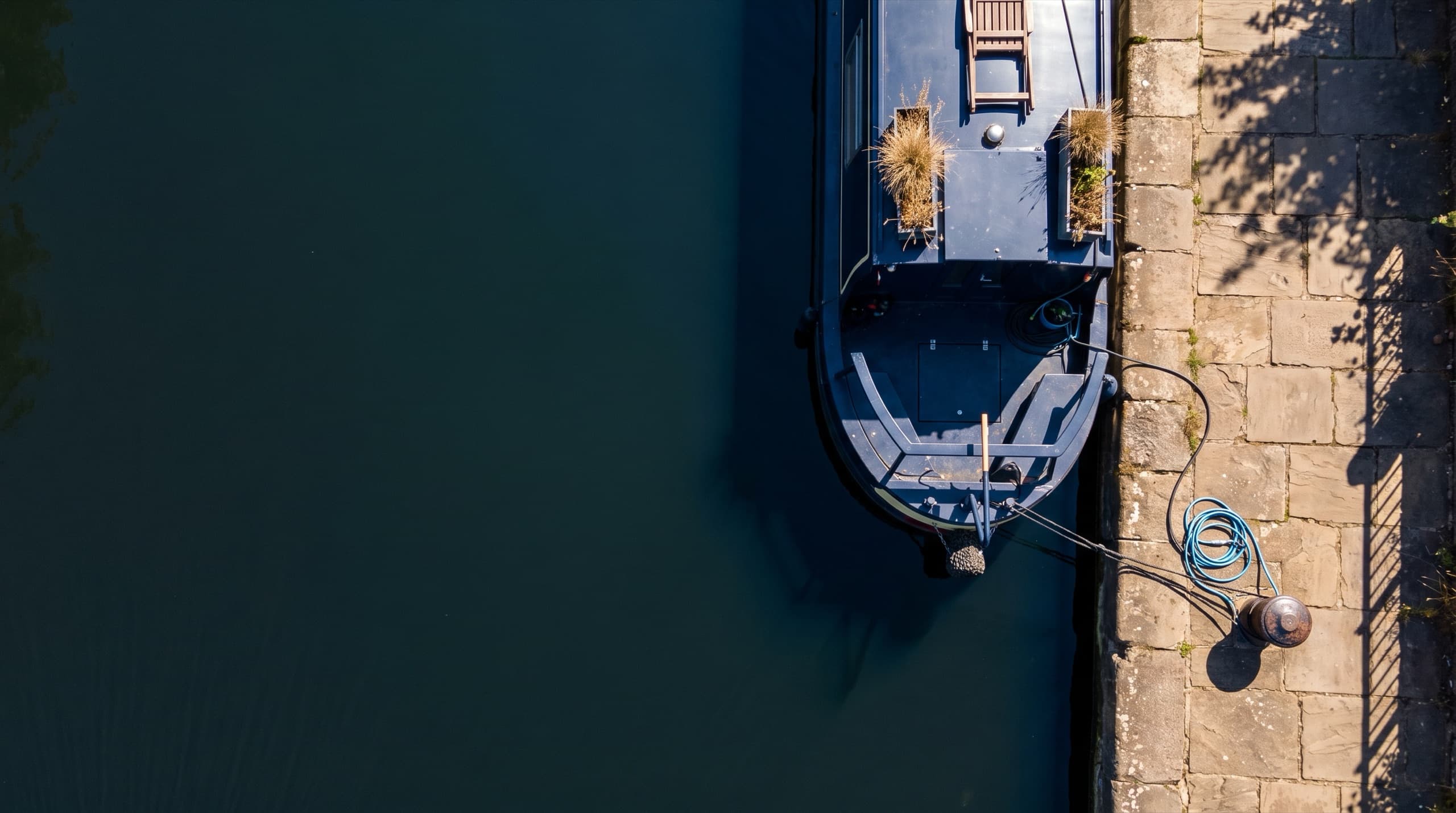 A widebeam houseboat moored against a stone quay on a still inland waterway in full daylight, shore power and water lines made up to a bollard on the towpath