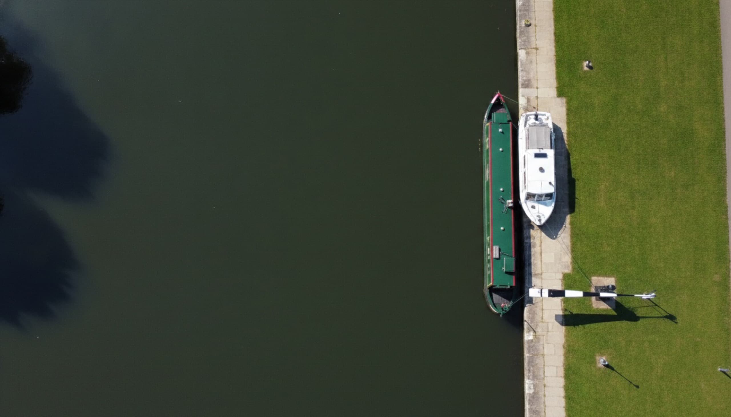 A steel narrowboat and a small white river cruiser moored bow to stern against a sunlit stone quay on an English inland navigation, seen from above in bright midday sunlight, with deep dark water filling the rest of the frame.