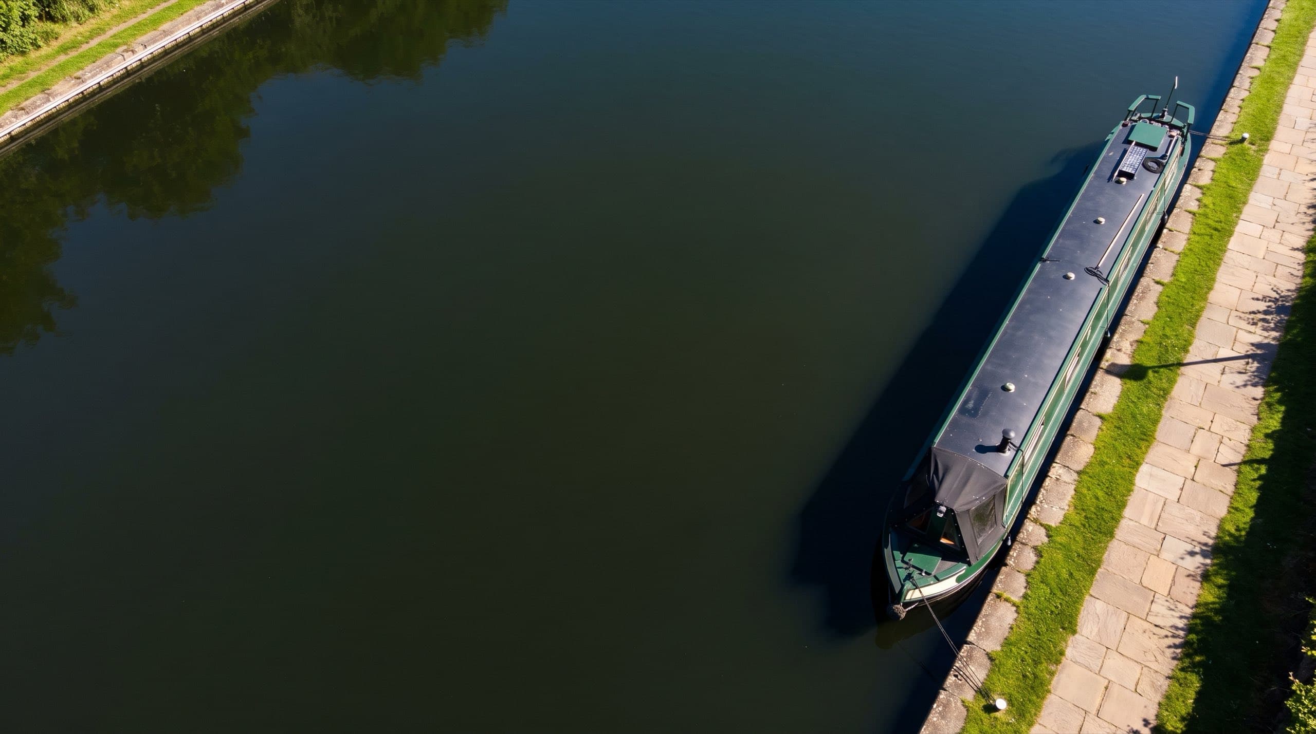 A moored steel narrowboat with a dark green hull alongside a sunlit stone towpath, still dark canal water filling the rest of the frame on a clear summer day