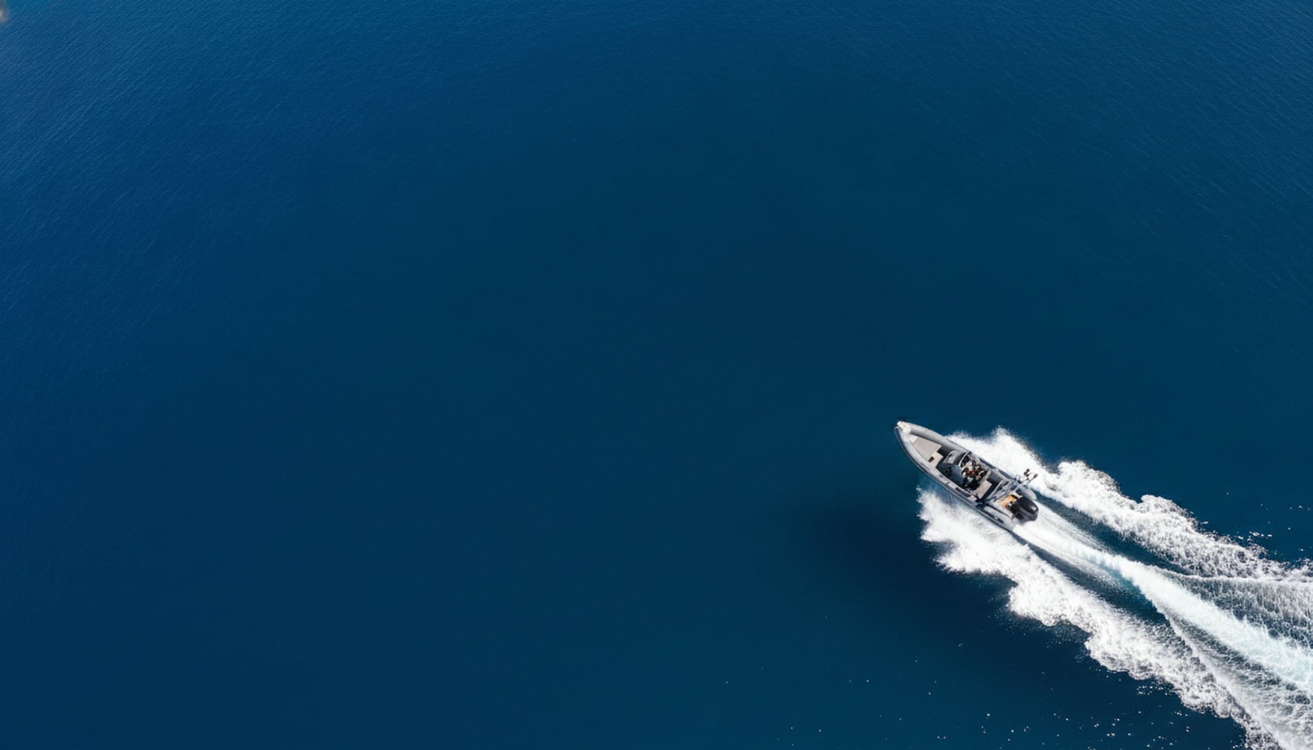 A grey rigid inflatable boat running at speed across deep blue open water in bright midday sun, throwing a white wake, seen from a high oblique aerial angle.