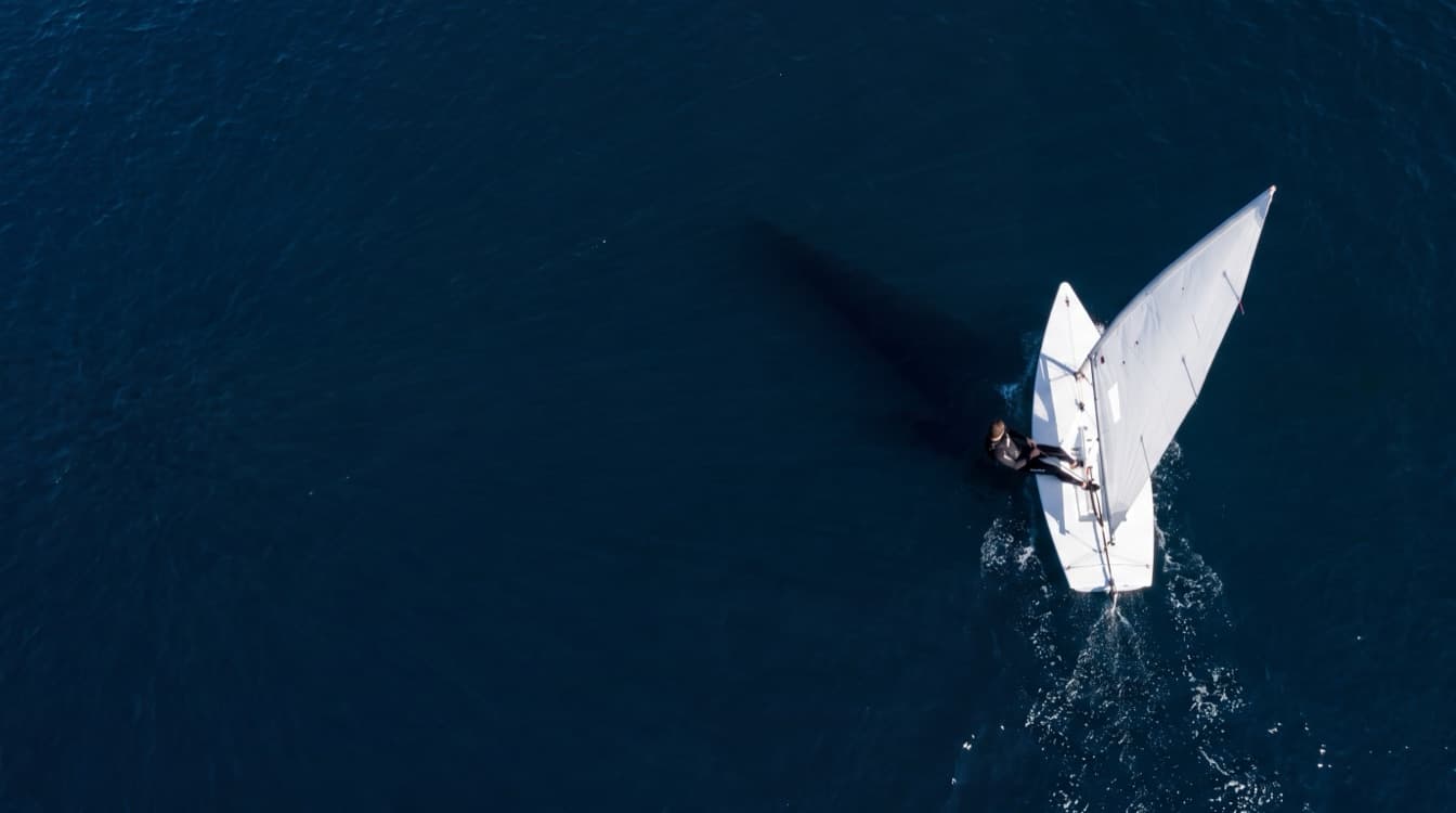 A single handed sailing dinghy heeling in bright sunshine on deep blue water off a low green headland