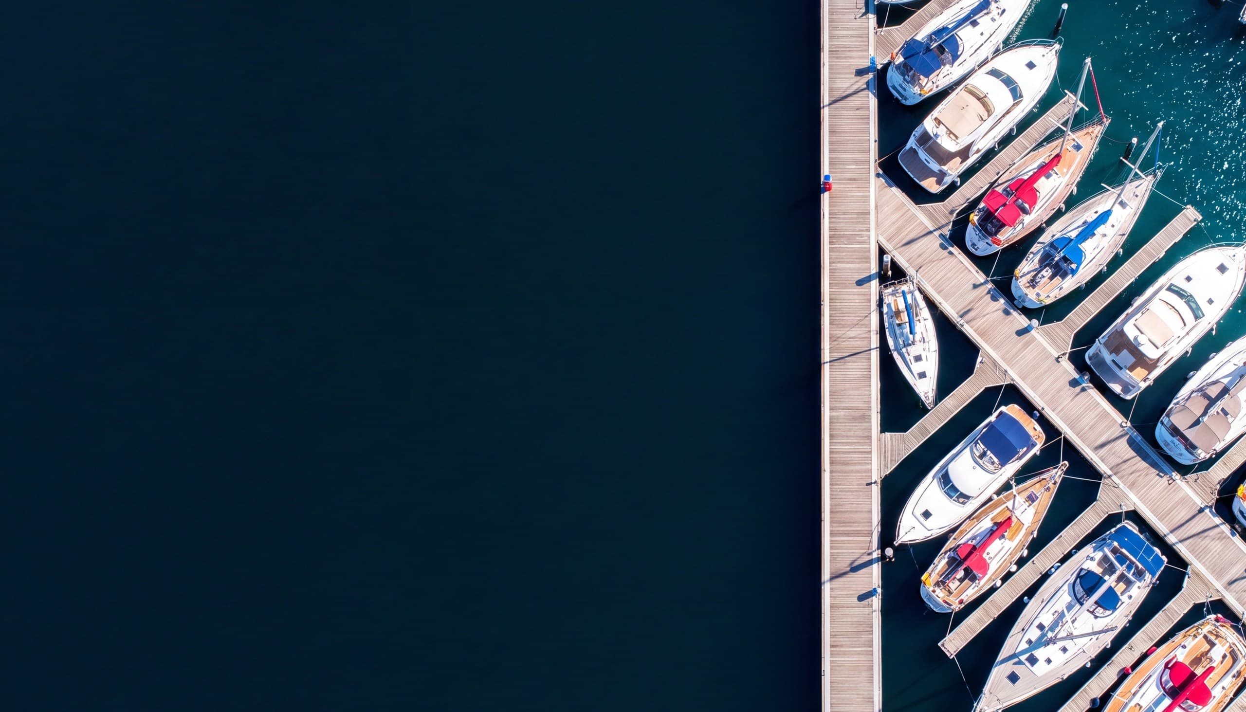 Aerial view of a sunlit marina, with sailing yachts and motor cruisers moored in their berths along timber pontoons beside deep open water