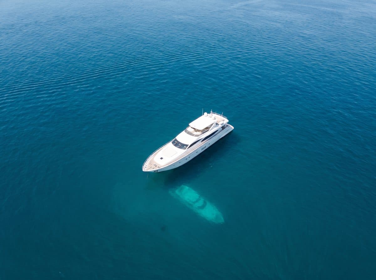 Uno yacht a motore bianco all'ancora da solo su acqua aperta blu intenso, visto dall'alto